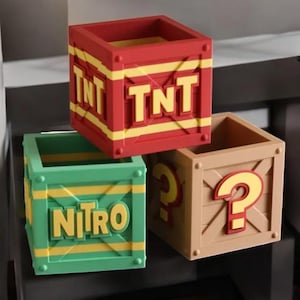 May include: Three colorful, square boxes with open tops. One is red with yellow accents and the letters "TNT". Another is green with yellow accents and the word "NITRO". The third is tan with a red and yellow question mark.