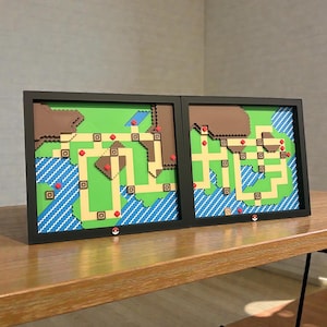 May include: Two framed pixel art maps, likely from a video game, sit side-by-side on a wooden surface. The maps feature green, tan, blue, brown, and red elements, with a black frame. A small red and white ball is at the bottom.