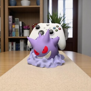 May include: A purple Gengar-themed controller holder with red eyes and a mischievous grin, holding a white Xbox controller. The holder is designed to resemble Gengar's form, resting on a cloud-like base. A fun accessory for gamers.