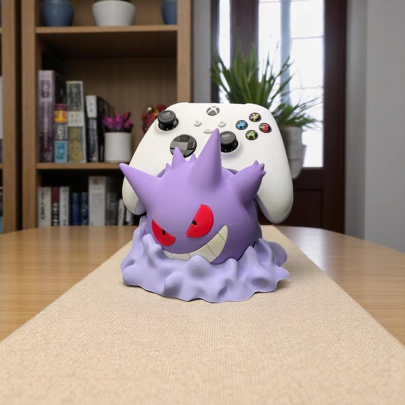 May include: A purple Gengar-themed controller holder with red eyes and a mischievous grin, holding a white Xbox controller. The holder is designed to resemble Gengar's form, resting on a cloud-like base. A fun accessory for gamers.