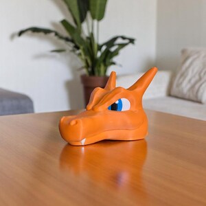 May include: An orange Charizard head figurine with blue and white eyes. The figurine is sitting on a wooden table. A potted plant is in the background.