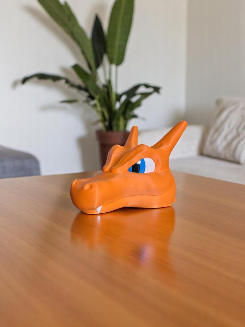 May include: An orange Charizard head figurine with blue and white eyes. The figurine is sitting on a wooden table. A potted plant is in the background.