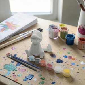 May include: A white, unpainted dinosaur figurine sits on a wooden surface, surrounded by paint supplies. Small paint pots in various colors, paintbrushes, and a book are also present. Splatters of paint in multiple colors cover the surface, suggesting a creative activity.
