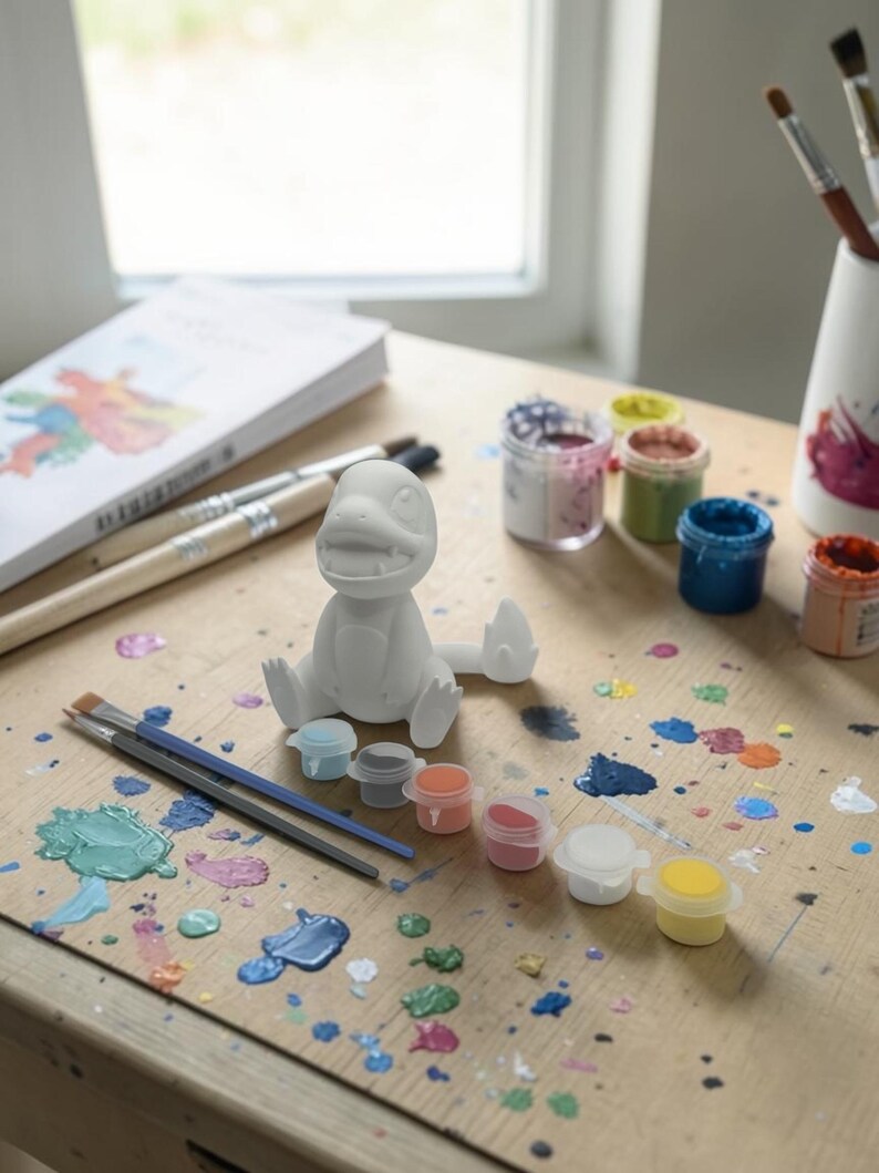 May include: A white, unpainted dinosaur figurine sits on a wooden surface, surrounded by paint supplies. Small paint pots in various colors, paintbrushes, and a book are also present. Splatters of paint in multiple colors cover the surface, suggesting a creative activity.