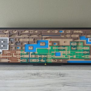 May include: A framed, layered artwork depicting a map with brown, green, blue, and white elements. The map is set against a neutral background and features a black frame. A gold triangle is at the bottom.