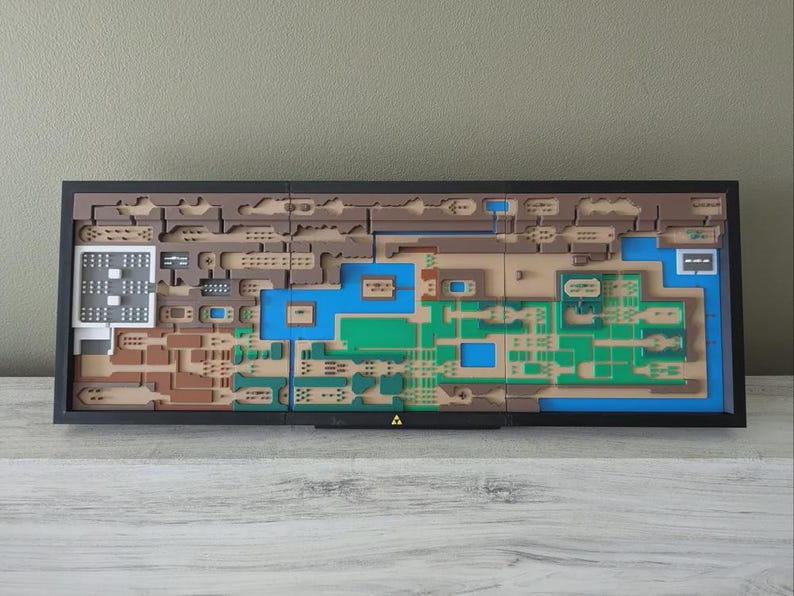May include: A framed, layered artwork depicting a map with brown, green, blue, and white elements. The map is set against a neutral background and features a black frame. A gold triangle is at the bottom.