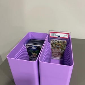 K&ouml;nnte beinhalten: Zwei lavendelfarbene Kartenaufbewahrungsboxen. Jede Box hat Trennw&auml;nde und enth&auml;lt Sammelkarten. Eine Box enth&auml;lt eine bewertete Karte in einer Schutzh&uuml;lle, die andere mehrere Karten.