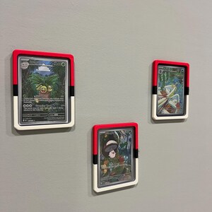 Puede incluir: Tres cartas coleccionables enmarcadas montadas en una pared gris. Cada marco tiene un borde rojo, un marco interior blanco y una pieza lateral negra. Las cartas presentan ilustraciones coloridas de personajes.