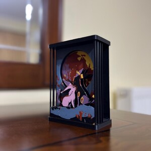 K&ouml;nnte beinhalten: Eine schwarze Holz-Schattenbox mit einer 3D-Szene von zwei rosa und violetten Comicfiguren, die vor einem roten und gelben Mond stehen. Die Figuren sind von einem dunkelblauen und violetten Hintergrund umgeben.
