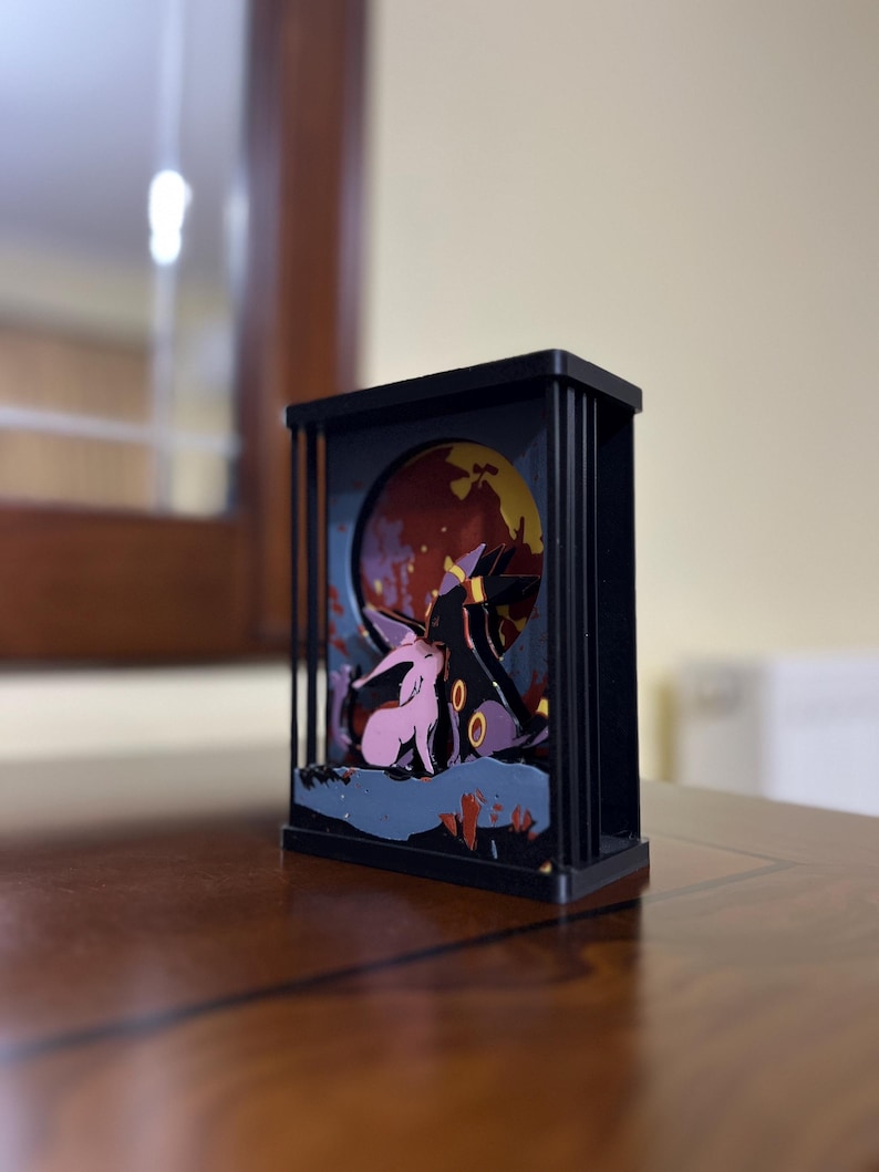 K&ouml;nnte beinhalten: Eine schwarze Holz-Schattenbox mit einer 3D-Szene von zwei rosa und violetten Comicfiguren, die vor einem roten und gelben Mond stehen. Die Figuren sind von einem dunkelblauen und violetten Hintergrund umgeben.