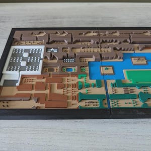May include: A multi-colored, 3D-printed map with a black frame. The map features various sections in shades of brown, green, blue, and white, with intricate details and textures. The overall design suggests a topographical or architectural model.