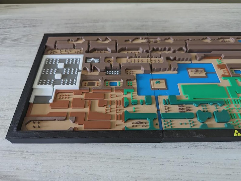May include: A multi-colored, 3D-printed map with a black frame. The map features various sections in shades of brown, green, blue, and white, with intricate details and textures. The overall design suggests a topographical or architectural model.