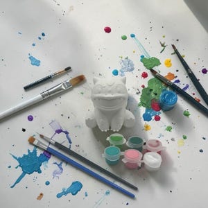 May include: A white ceramic figure of a cartoon character, surrounded by paintbrushes and small paint pots. Splatters of blue, green, pink, and yellow paint are scattered across the white surface, suggesting a creative activity.