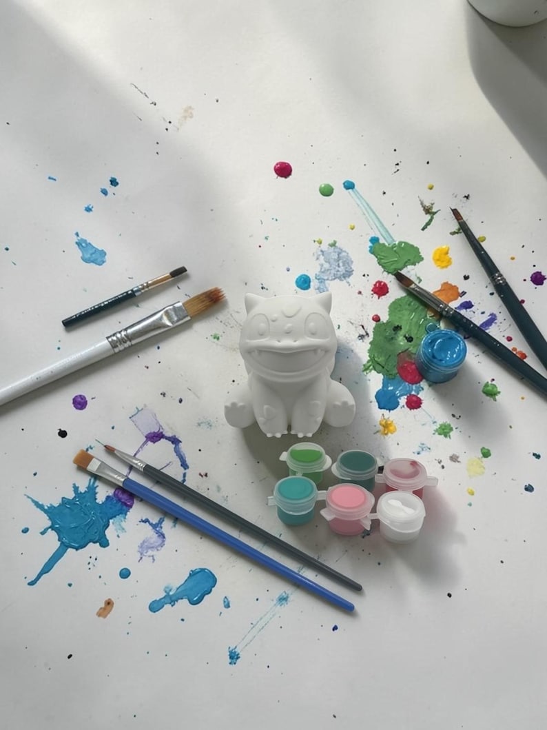 May include: A white ceramic figure of a cartoon character, surrounded by paintbrushes and small paint pots. Splatters of blue, green, pink, and yellow paint are scattered across the white surface, suggesting a creative activity.
