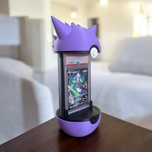 以下が含まれることがあります： 紫と黒のポケモンカードディスプレイスタンド。スタンドはゲンガーにインスパイアされたデザインで、中にグレードカードが見えます。スタンドは木製の表面にあります。