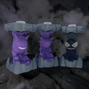 May include: Three stylized figures of Pokemon characters Gengar, Haunter, and Gastly. Each figure is purple or black, and sits atop a gray, geometric base. The background is dark with a smoky effect, creating a spooky atmosphere.