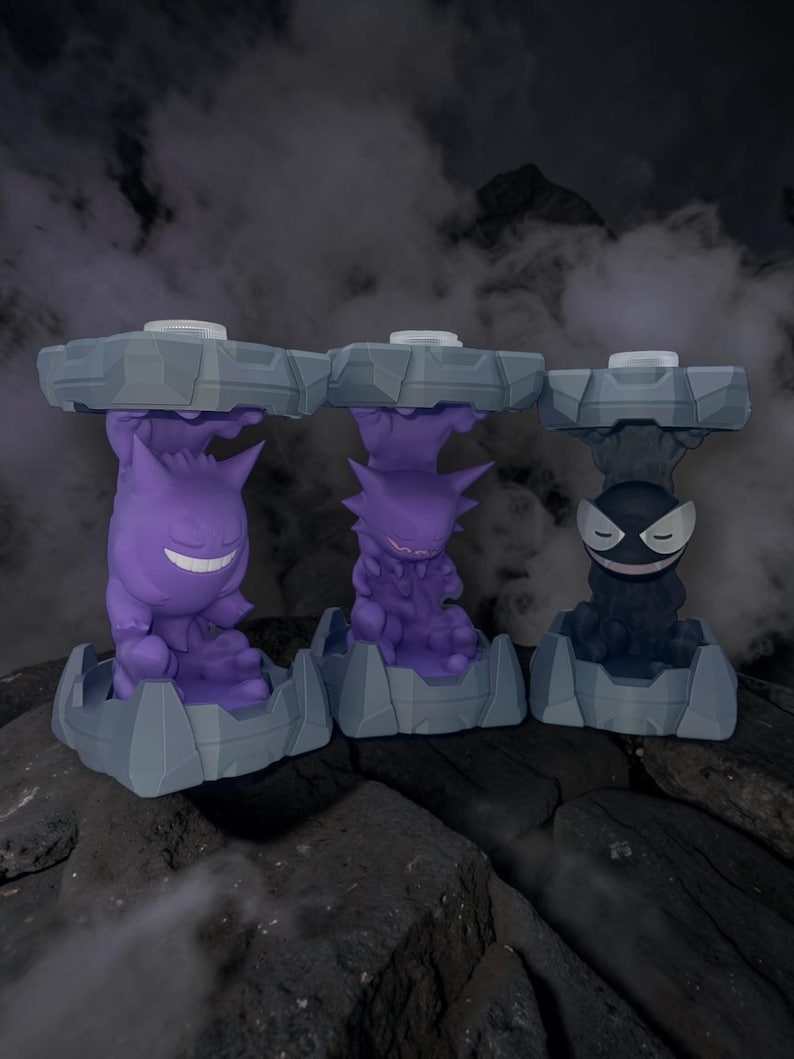 May include: Three stylized figures of Pokemon characters Gengar, Haunter, and Gastly. Each figure is purple or black, and sits atop a gray, geometric base. The background is dark with a smoky effect, creating a spooky atmosphere.