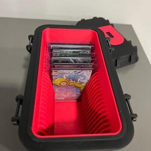 K&ouml;nnte beinhalten: Schwarze und rote Kartenaufbewahrungsbox, ge&ouml;ffnet, um mehrere Sammelkarten zu zeigen. Der Innenraum ist rot mit vertikalen Trennw&auml;nden. Der Deckel ist teilweise sichtbar, mit einem roten Buchstaben "P" darauf. Die Box dient zum Schutz und zur Organisation von Sammelkarten.