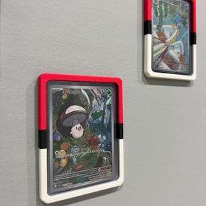 Puede incluir: Dos cartas coleccionables enmarcadas est&aacute;n montadas en una pared gris. Los marcos son rectangulares con una parte superior roja, una franja negra y una parte inferior blanca. Las cartas presentan ilustraciones coloridas de personajes y texto.