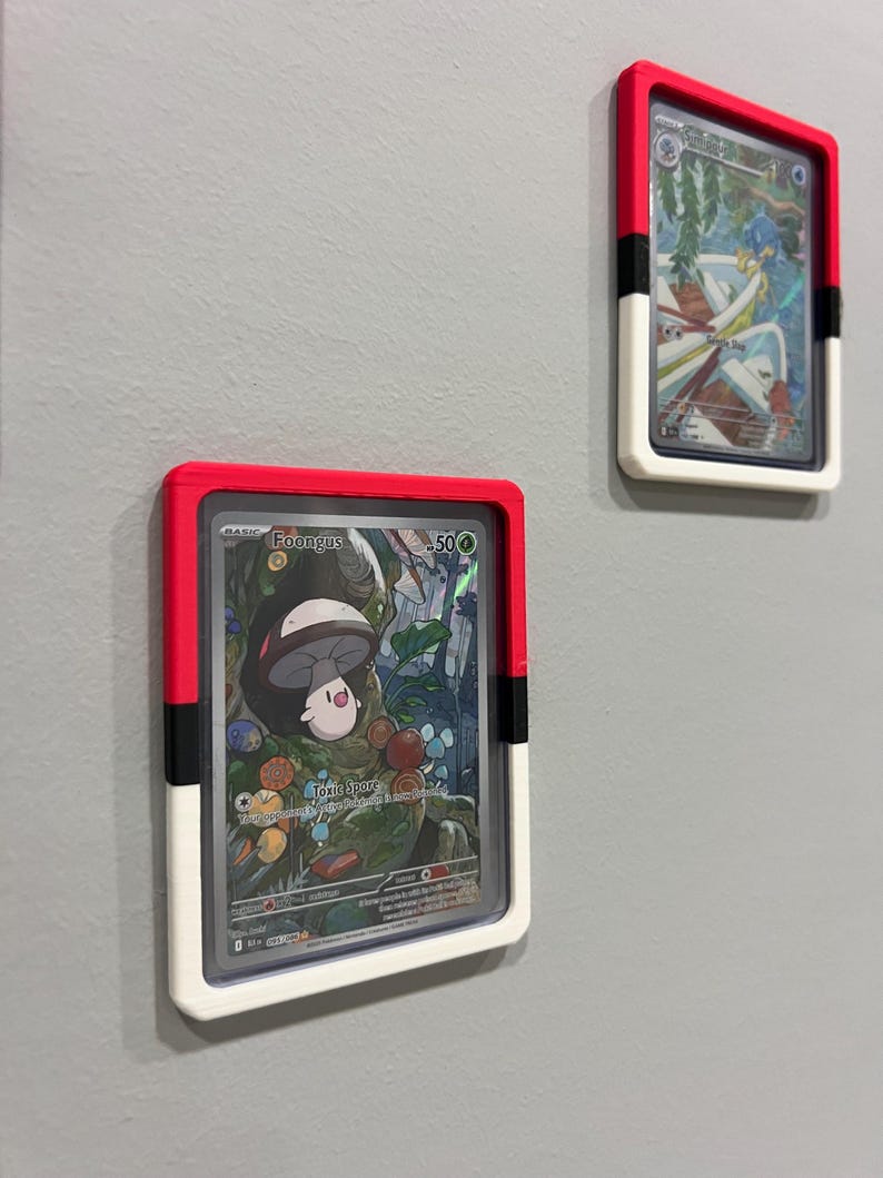 Puede incluir: Dos cartas coleccionables enmarcadas est&aacute;n montadas en una pared gris. Los marcos son rectangulares con una parte superior roja, una franja negra y una parte inferior blanca. Las cartas presentan ilustraciones coloridas de personajes y texto.