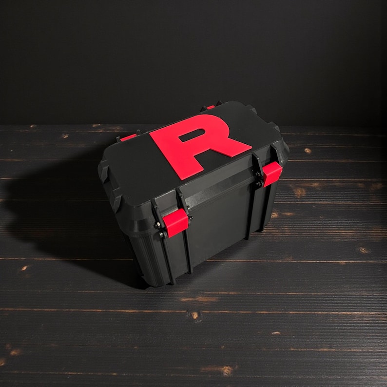 K&ouml;nnte beinhalten: Schwarze Aufbewahrungsbox mit einem gro&szlig;en roten "R" auf dem Deckel. Die Box hat rote Verschl&uuml;sse und Scharniere. Die Box steht auf einer dunklen Holzoberfl&auml;che.