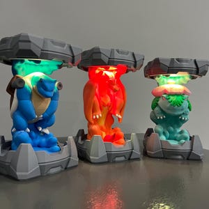 May include: Three colorful character lamps. The lamps feature a blue turtle-like creature, an orange dragon-like creature, and a green plant-like creature. Each lamp has a gray base and a gray top with a light shining from underneath.