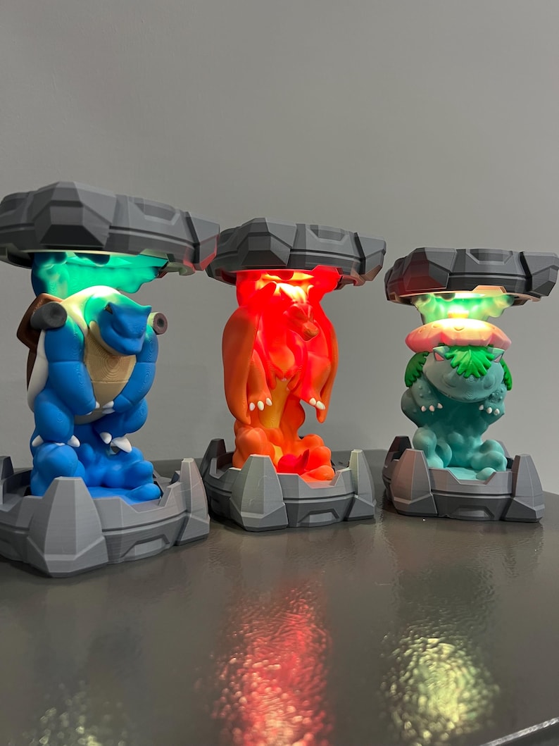 May include: Three colorful character lamps. The lamps feature a blue turtle-like creature, an orange dragon-like creature, and a green plant-like creature. Each lamp has a gray base and a gray top with a light shining from underneath.