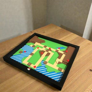 May include: A framed, pixelated map featuring a green landscape with brown hills, blue water, and a network of beige paths. Red accents mark key locations. A white and red Pokeball is on the frame. The artwork is displayed on a wooden table.