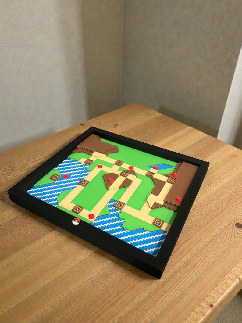May include: A framed, pixelated map featuring a green landscape with brown hills, blue water, and a network of beige paths. Red accents mark key locations. A white and red Pokeball is on the frame. The artwork is displayed on a wooden table.