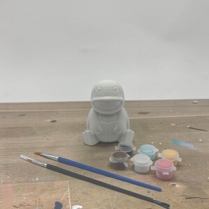 May include: A white, unpainted ceramic figure of a cartoon character, with paint pots and paintbrushes. The figure is sitting on a wooden surface. The paint pots are small and contain various colors.