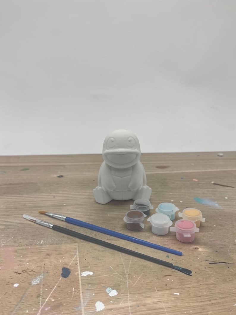 May include: A white, unpainted ceramic figure of a cartoon character, with paint pots and paintbrushes. The figure is sitting on a wooden surface. The paint pots are small and contain various colors.