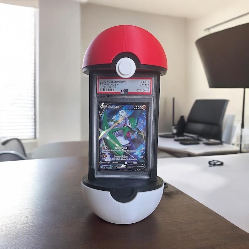 Op de afbeelding: Een rood en wit Pok&eacute; Ball-vormig display met een Pok&eacute;mon-ruilkaart. De kaart zit in een doorzichtige plastic houder en heeft een Gallade V-kaart. Het display staat op een houten tafel.