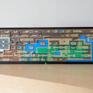 Op de afbeelding: Een rechthoekige, veelkleurige 3D-kaart van een videogame-wereld, geplaatst in een zwart frame. De kaart bevat bruine, groene, blauwe en witte secties, met ingewikkelde details die verschillende locaties en paden vertegenwoordigen. De kaart wordt weergegeven op een lichtgekleurd houten oppervlak.