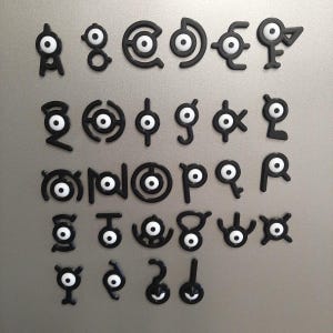 May include: A set of black Unown letter magnets with white circular centres, displayed on a silver surface. The magnets are in diverse shapes, representing the alphabet from a popular franchise.