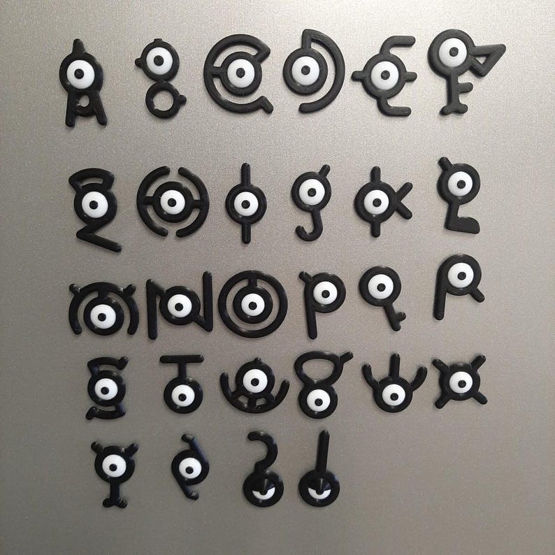 May include: A set of black Unown letter magnets with white circular centres, displayed on a silver surface. The magnets are in diverse shapes, representing the alphabet from a popular franchise.