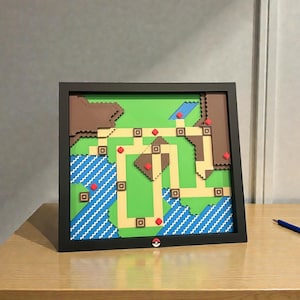 May include: A framed, layered artwork depicting a colorful map with green, brown, blue, and tan sections. The map features a grid-like pattern with red and brown accents, and a Pok&eacute; Ball logo is visible. The artwork is displayed on a wooden table.