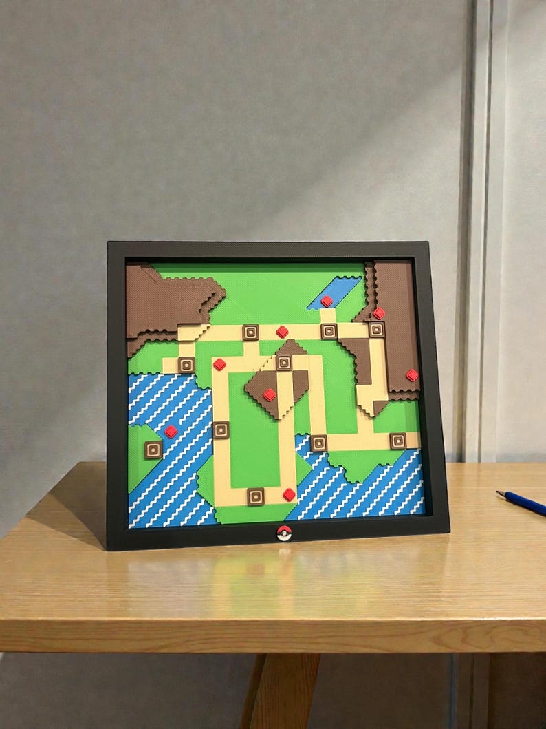 May include: A framed, layered artwork depicting a colorful map with green, brown, blue, and tan sections. The map features a grid-like pattern with red and brown accents, and a Pok&eacute; Ball logo is visible. The artwork is displayed on a wooden table.