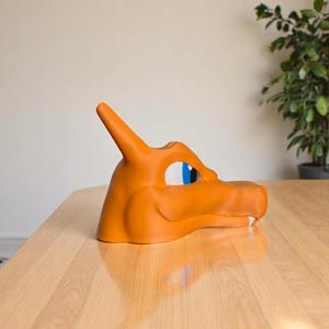 May include: An orange Charizard mask, a character from the Pok&eacute;mon franchise, is displayed on a wooden table. The mask features blue and white eye details and a horn. The mask is made of a smooth material.