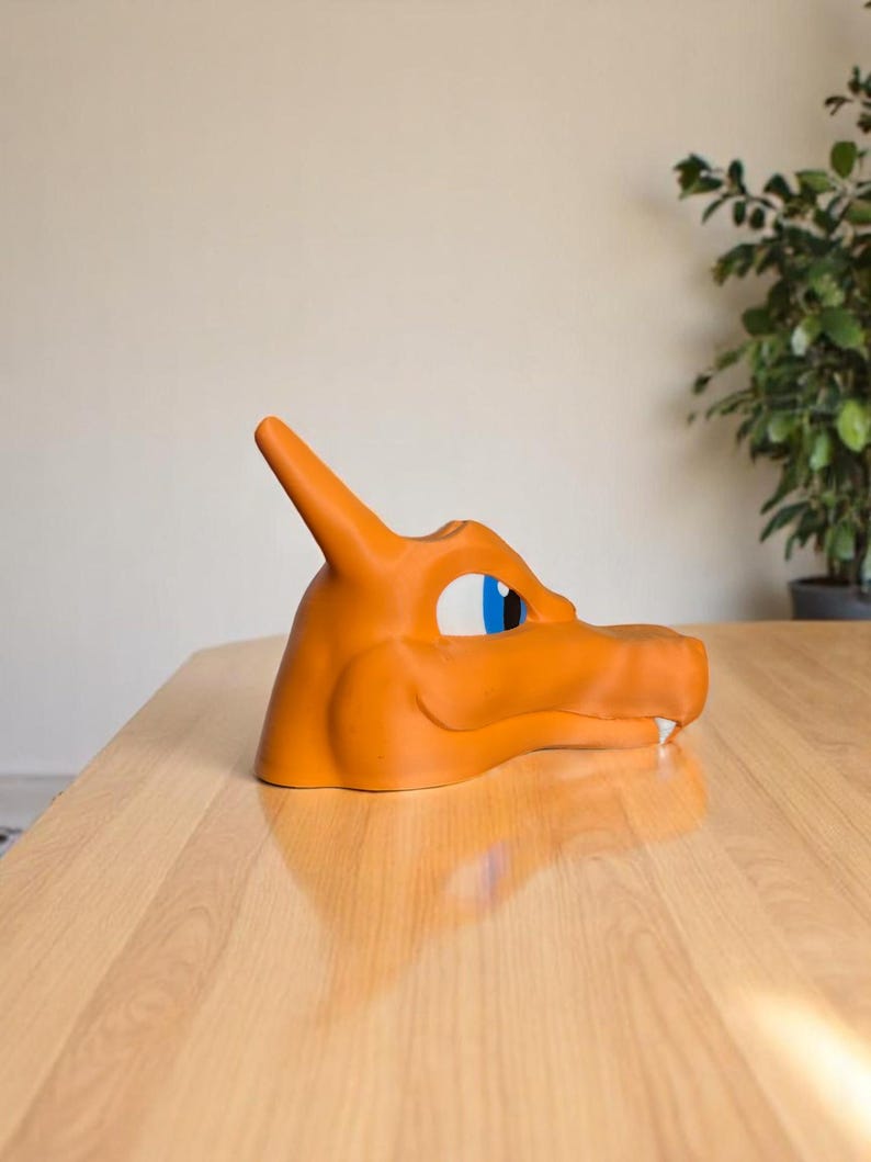 May include: An orange Charizard mask, a character from the Pok&eacute;mon franchise, is displayed on a wooden table. The mask features blue and white eye details and a horn. The mask is made of a smooth material.