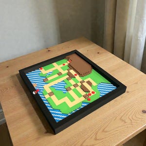 May include: A framed, pixelated map featuring green land, blue water, and brown structures. Red markers indicate locations. The map is set within a black frame, creating a 3D effect. The overall design is reminiscent of a video game map.