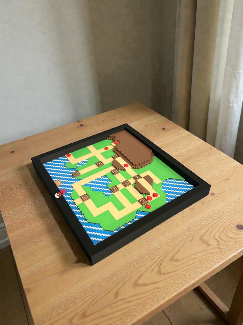 May include: A framed, pixelated map featuring green land, blue water, and brown structures. Red markers indicate locations. The map is set within a black frame, creating a 3D effect. The overall design is reminiscent of a video game map.