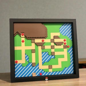 May include: A framed map of a video game world, featuring a green landscape with tan pathways, brown mountains, and blue water. Red markers and a small Pok&eacute; Ball are included. The frame is black.