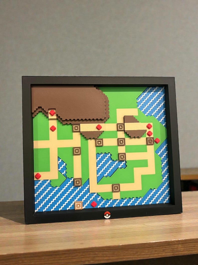 May include: A framed map of a video game world, featuring a green landscape with tan pathways, brown mountains, and blue water. Red markers and a small Pok&eacute; Ball are included. The frame is black.