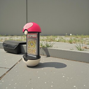 Puede incluir: Un expositor rosa y blanco con forma de Pok&eacute; Ball, que contiene una carta coleccionable calificada. El expositor est&aacute; sobre una superficie gris con un estuche negro en el fondo. La carta es visible dentro de una funda protectora transparente.