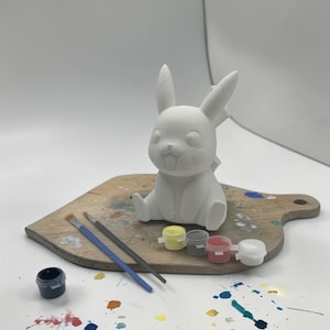May include: A white ceramic Pikachu figurine, ready to be painted, sits on a wooden cutting board. Paintbrushes and small paint pots in various colors are arranged nearby. Splatters of paint add a colorful accent to the scene.