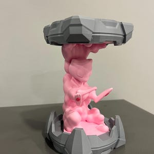May include: A 3D-printed sculpture featuring a pink figure of Mewtwo, supported by a gray base and a top structure. The sculpture has a detailed design with geometric shapes and a smooth finish. The figure is in a dynamic pose, with a red accent on its hand.