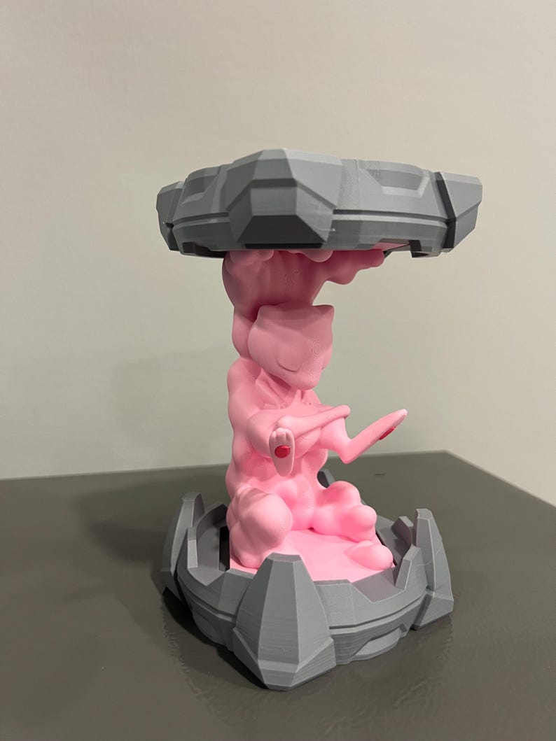 May include: A 3D-printed sculpture featuring a pink figure of Mewtwo, supported by a gray base and a top structure. The sculpture has a detailed design with geometric shapes and a smooth finish. The figure is in a dynamic pose, with a red accent on its hand.