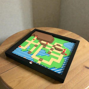 May include: A framed, colorful 3D map of a video game world. The map features green land, blue water, and brown mountains, with a black frame. A red and white Pokeball is on the frame.