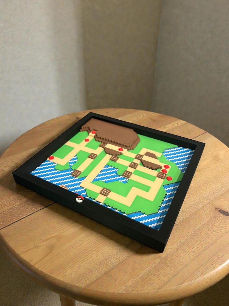 May include: A framed, colorful 3D map of a video game world. The map features green land, blue water, and brown mountains, with a black frame. A red and white Pokeball is on the frame.