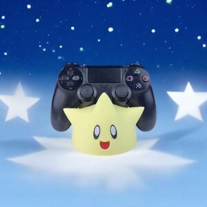 K&ouml;nnte beinhalten: Ein schwarzer Videospiel-Controller sitzt auf einem gelben sternf&ouml;rmigen St&auml;nder mit einem l&auml;chelnden Gesicht. Der Hintergrund ist ein blauer Himmel mit wei&szlig;en Sternen.
